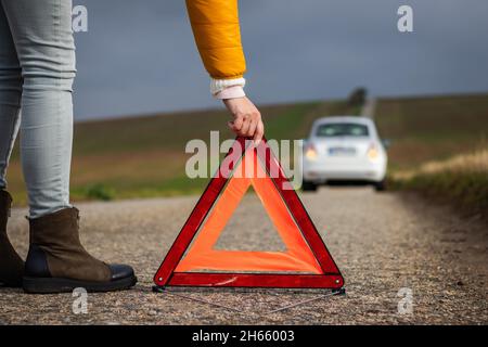 Frau, die ein Warndreieck auf die Straße legt, weil das Auto kaputt ist. Rotes Warnschild vor Ankunft des Pannenassistierten. Stockfoto