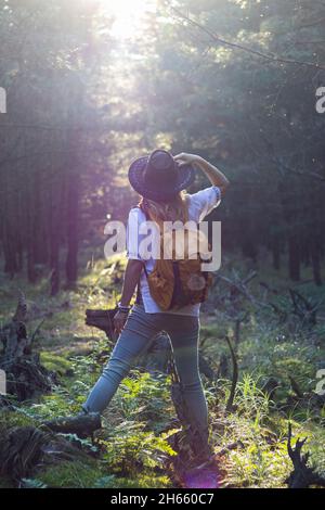 Wanderfrau mit Hut und Rucksack und Blick in die Sonne im Wald. Weibliche Reisende steht im Sonnenstrahl am Wald. Genuss der Natur Stockfoto