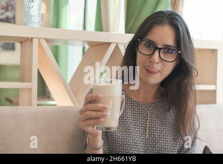 asiatische Frau trägt Brillen trinken Latte Kaffee und zeigt eine Zunge. Verbringen Sie Zeit im Café, Entspannung und Spaß haben. Stockfoto