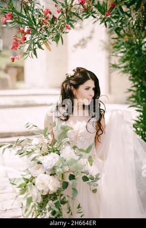 Die Braut steht unter einem blühenden Oleander mit einem Bouquet aus weißen Rosen, Pfingstrosen, Delphinien und Eukalyptuszweigen in ihren Händen Stockfoto