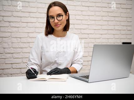 Schöne selbstbewusste Frau Arzt, Allgemeinmediziner in weißer medizinischer Uniform sitzen am Schreibtisch im Krankenhaus schriftlich und Beratung Patient online Stockfoto