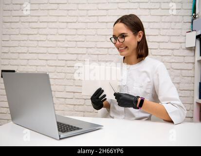 Die charmante junge Ärztin konsultiert online, während sie am Tisch im Krankenhaus sitzt und einem Patienten beim Lapto ein Notizbuch zeigt Stockfoto