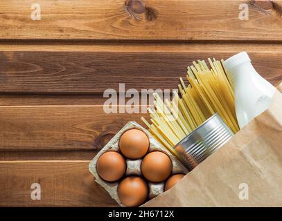 Papiertüte mit Lebensmitteln Krisenlager für Quarantäneisolation auf Holzhintergrund. Eier, Milch, Konserven, Pasta. Lieferung von Lebensmitteln Stockfoto