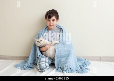Ein Junge im grauen Schlafanzug sitzt auf einer blauen Strickdecke mit einer schönen weißen Katze auf dem Boden, Stockfoto