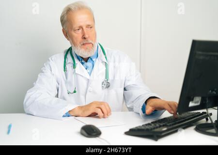 Porträt eines fokussierten reiferen männlichen Arztes mit weißem Mantel und Stethoskop, der am Desktop-Computer am Schreibtisch im medizinischen Büro sitzt. Stockfoto