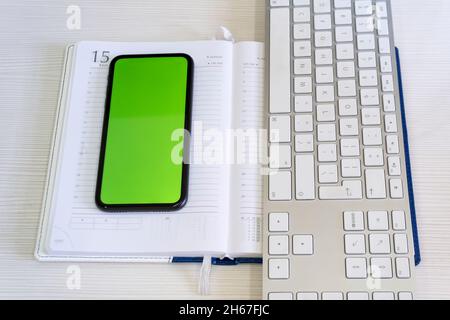 Ein Smartphone mit grünem Bildschirm liegt auf einem Notebook, eine Computertastatur ist daneben, die digitale Welt steht auf einer Papierwelt. Stockfoto
