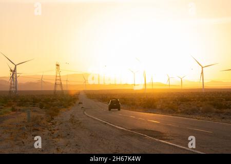 Auto, das mit Windmühlen und Strommasten in den Sonnenuntergang fährt Stockfoto