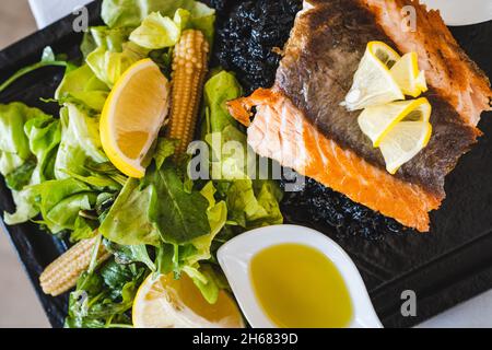 Geröstetes Lachsfilet auf schwarzem Teller mit Zitrone und Salat Draufsicht Stockfoto