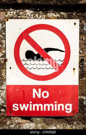 Rotes Warnschild ohne Schwimmen, das an einer Backsteinmauer neben einem Fluss befestigt ist Stockfoto