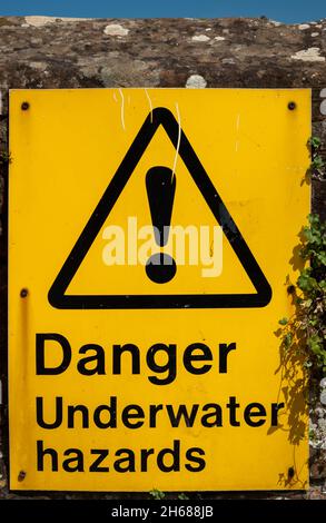 Ein gelbes Warnschild für Gefahren unter Wasser, das an einer Ziegelmauer neben einem Fluss befestigt ist Stockfoto