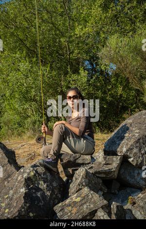 Eine junge asiatische Frau, die auf einem Felsen im Fluss sitzt, während sie Fliegenfischen geht Stockfoto