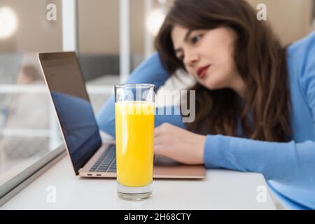 Vor einem Laptop sitzt eine attraktive junge Frau mit einem Glas Saft. Stockfoto