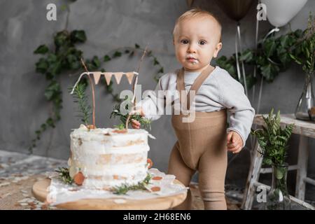 Kid boy Kuchen essen an seinem ersten Geburtstag, ein Jahr altes Kind leckeren Kuchen, dekoriert Hintergrund in natürlichen Stil, Nahaufnahme von Jungen lecken Creme Stockfoto