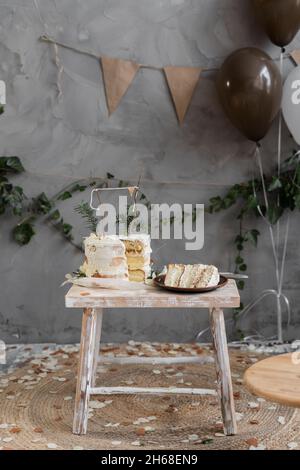 Dekoriert mit Cremekuchen zum ersten Geburtstag, stehend auf einem kleinen Holztisch, einem geschnittenen Stück Kuchen auf einem Teller, einem Raum mit handgefertigten Dekorationen. Stockfoto