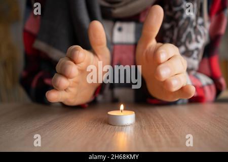 Nahaufnahme der männlichen Hände, die sich in der Nähe einer brennenden Kerze aufwärmen, die auf dem Tisch steht. Konzept der Probleme mit der Heizung, Kälte zu Hause im Winter. Stockfoto