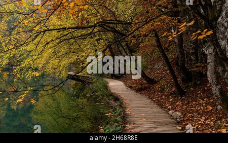 Eine rustikale Holzpromenade, die entlang des Ufers eines malerischen Bergsees mit Bäumen und Laub in intensiven Herbstfarben führt Stockfoto