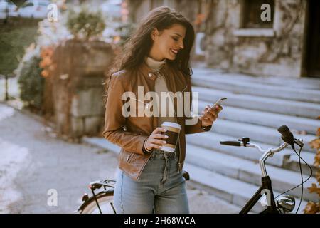 Hübsche junge Frau mit Handy trinkt am Herbsttag Kaffee, um mit dem Fahrrad zu fahren Stockfoto