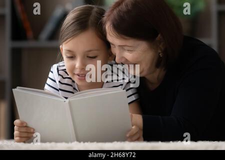 Lächelnde reife Großmutter mit einem kleinen Mädchen, das zusammen Buch liest Stockfoto