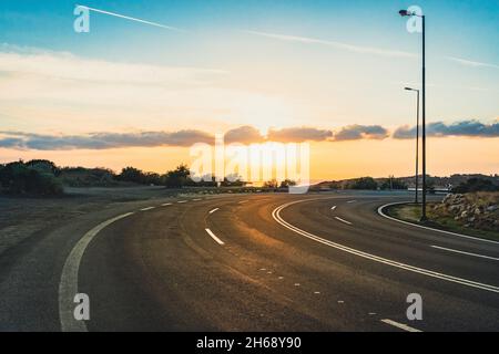 Landstraße mit Kurve, aber keine Autos bei Sonnenuntergang. Asphaltstraße auf einem kleinen Hügel mit Meerblick im Hintergrund. Stockfoto