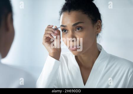 Schöne junge afroamerikanische Frau im Bademantel mit einer Pinzette, Stockfoto