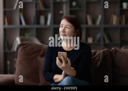 Verträumte reife Frau mit modernem Smartphone, die zur Seite schaut Stockfoto