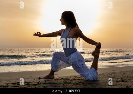 Attraktive junge Frau praktiziert Yoga am Meer bei Sonnenuntergang. Frau trainiert die einbeinige King Pigeon Übung, Eka Pada Rajakapotasana Yoga-Pose. Stockfoto