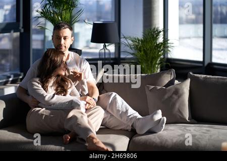 Ein hübscher kaukasischer Mann und eine schöne Frau sitzen in Umarmungen, während sie sich auf der bequemen Couch ausruhen. Tageslichter durch Panoramafenster in einem modernen Apartment. Entspannung zu Hause. Stockfoto