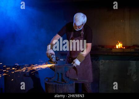 Grauhaariger Mann in Sicherheitsschürze und Handschuhe, die Metall in der Werkstatt mit Winkelschleifer schweißen. Kaukasischer Handwerker formt Stahl am Arbeitsplatz. Stockfoto