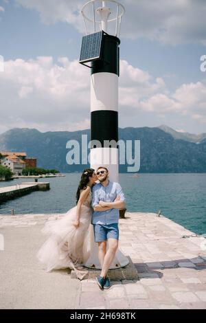 Die Braut und der Bräutigam umarmen sich am Pier in der Nähe des Leuchtturms in der Bucht von Kotor, die Braut küsst den Bräutigam Stockfoto