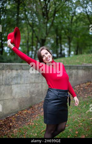 Lächelnde weiße Frau mit langen dunklen Haaren in leuchtendem Rot in der Nähe einer Steinmauer im Herbstpark, die ihren Hut in der rechten Hand hält Stockfoto