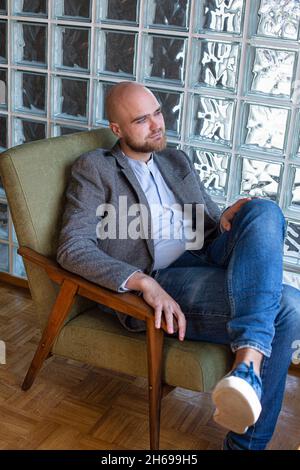 Weißer europäischer Glatze mit Bart und grauen Augen in der Jacke und Jeans auf einem alten Stuhl Stockfoto