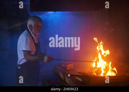 Reifer kaukasischer Handwerker mit Schutzhandschuhen und Schürze, die Stahl im Schmiedeofen anhärte. Professioneller Schmied, der Industriewerkzeuge verwendet, während er mit geschmolzenem Metall arbeitet. Stockfoto