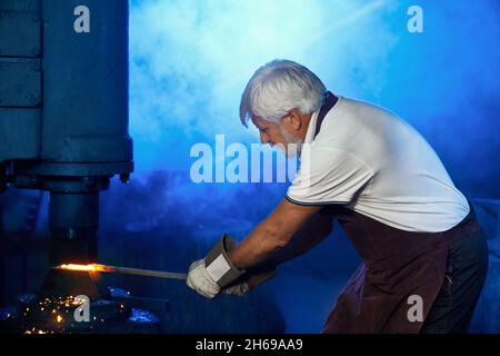 Starker reifer Schmied in Sicherheitsschürze und Handschuhe, die erhitzten Stahl unter der Pressenmaschine biegen. Kaukasischer Handwerker, der am Arbeitsplatz geschmolzenes Metall verarbeitet. Stockfoto