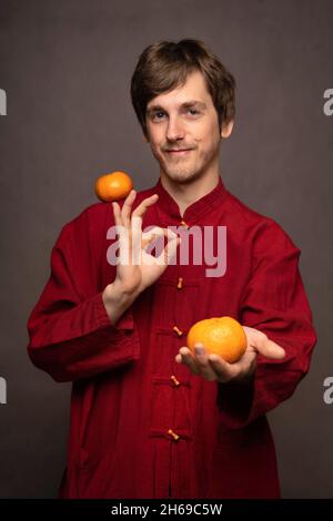 Junger, hübscher, großer, schlanker weißer Mann mit braunem Haar, Mandarine auf der Schulter und Orange in der Hand in einem roten Hemd auf grauem Hintergrund Stockfoto