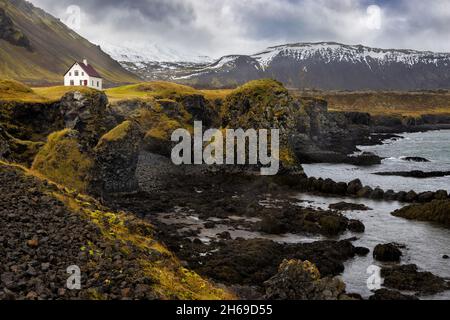 Arnarstapi, Island. Ein Blick auf die zerklüftete Küste mit einem einsamen Haus. Herbstfarben mit schneebedeckten Bergen im Hintergrund. Snaefellsnes Stockfoto