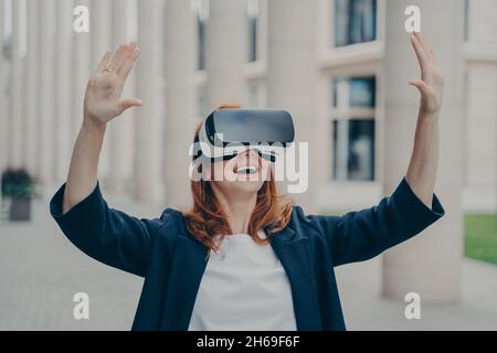 Beeindruckte Ingwer-Geschäftsfrau testet Virtual Reality, indem sie ein mobiles VR-Headset im Freien verwendet Stockfoto