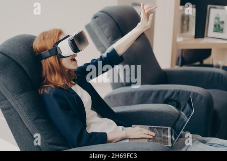 Aufgeregt Frau in VR-Headset und Laptop, berühren Luft mit der Hand, sitzen im Sessel im Café Stockfoto