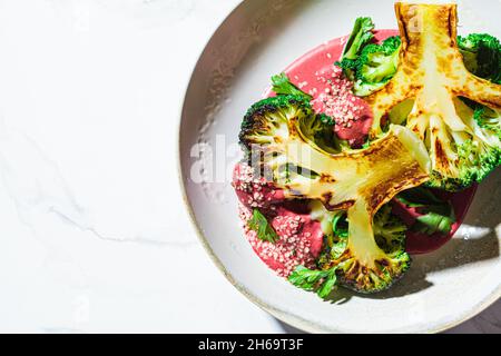 Gegrilltes Brokkoli-Steak mit rosafarbener Rote Beete-Sauce und Hanfsamen in einer weißen Schüssel, Hintergrund aus weißem Marmor, Kopierraum. Gesundes veganes Lebensmittelkonzept. Stockfoto