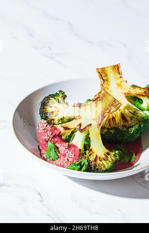 Gegrilltes Brokkoli-Steak mit rosafarbener Rote Beete-Sauce und Hanfsamen in einer weißen Schüssel, Hintergrund aus weißem Marmor, Kopierraum. Gesundes veganes Lebensmittelkonzept. Stockfoto