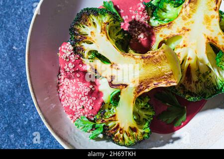 Gegrilltes Brokkoli-Steak mit rosa Rote-Bete-Sauce und Hanfsamen in weißer Schüssel, blauer Hintergrund, Nahaufnahme. Gesundes veganes Lebensmittelkonzept. Stockfoto