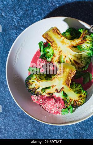 Gegrilltes Brokkoli-Steak mit rosa Rote-Bete-Sauce und Hanfsamen in weißer Schüssel, blauer Hintergrund, Nahaufnahme. Gesundes veganes Lebensmittelkonzept. Stockfoto