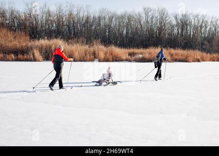 Familie auf Winterspaziergang am sonnigen Tag. Junge Frau, Mann und Kind fahren auf gefrorenem Fluss vor WaldHintergrund Ski, Mann reitet Kind auf Kindern Stockfoto