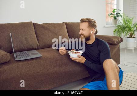 Ein fitter Mann isst, entspannt sich und schaut sich nach dem Fitnesstraining zu Hause Videos auf dem Laptop an Stockfoto