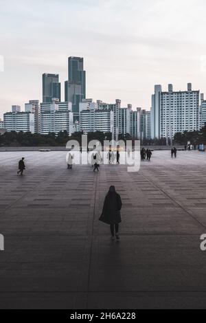 Junge Frau im schwarzen Mantel geht auf der Straße in der Metropole. Wolkenkratzer im Hintergrund. Menschen in schwarzen Kleidern gehen auf der Megapolis-Straße Stockfoto