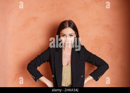 Business erfolgreiche Brünette Frau in goldenem Kleid und schwarzer Jacke posiert im Studio. Ein süßes kaukasisches Mädchen mit braunen Augen schaut auf die Kamera und lächelt Stockfoto