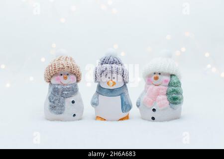 Dekorative Figuren im Weihnachtsstil. Die Statuette eines Pinguins und zweier Schneemänner in einem gestrickten Hut auf weißem Hintergrund. Festliche Einrichtung, warmes Bokeh Stockfoto