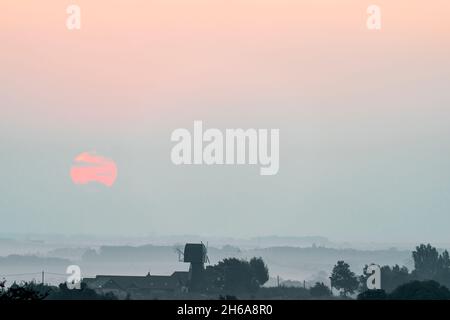 Sonnenaufgang in Kent, England. Eine teilweise verborgene Sonne, die über einer Wolkenschicht aufgeht, die kaum durch den Nebel, der den Boden bedeckt, gesehen wird. Im Vordergrund Bäume ein Stockfoto