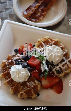 Leckere belgische Waffeln mit frischer Schlagsahne und Obst zusammen mit Speck auf der Seite Stockfoto