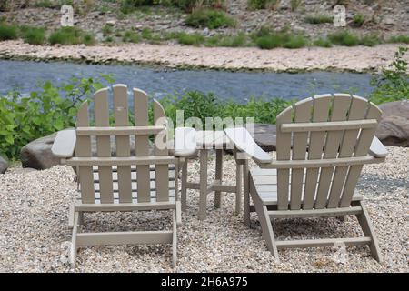 Zwei graue Adirondack-Stühle mit Blick auf einen fließenden Fluss mit felsiger Küste Stockfoto