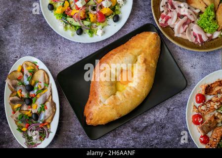 Draufsicht auf Khachapuri Gericht mit Käse und Ei in Adjarian mit Caesar und griechischen Salaten mit Kartoffeln und Hering auf dem Tisch Stockfoto